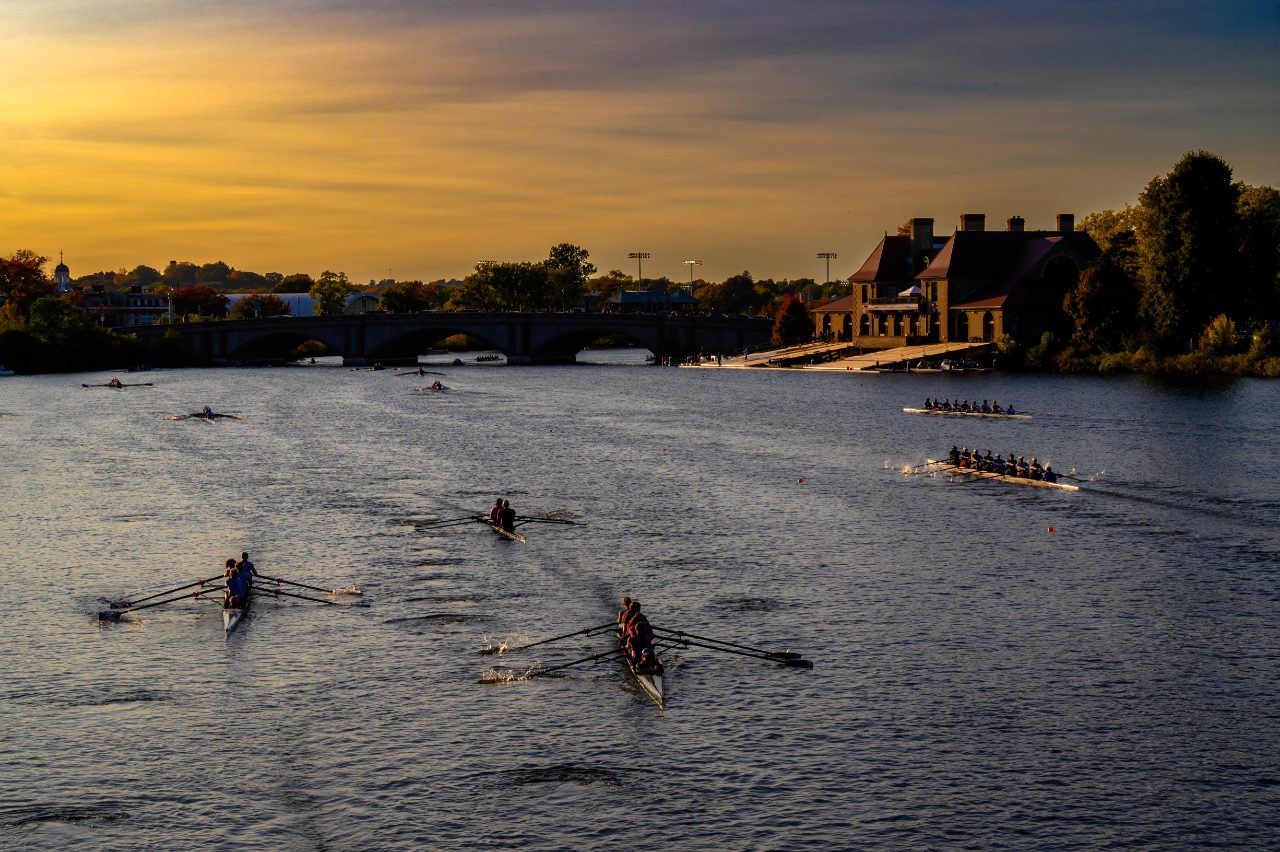 Crews racing on the Charles River at sunset, with a boathouse on the far bank